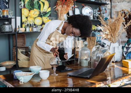 Pretty black woman in apron creating online catalogue using wireless laptop. African saleswoman in eyeglasses taking picture of wine glass in shop on modern smartphone. Stock Photo