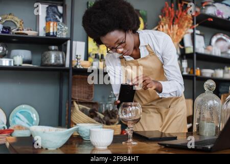 Close up of African saleswoman in eyeglasses taking picture of decorations and glass in shop on modern smartphone, creating online catalogue using wireless laptop. Stock Photo