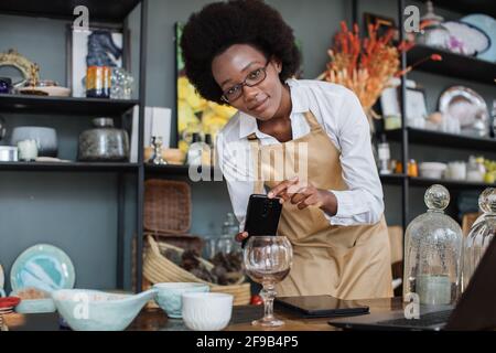 Close up of African saleswoman in eyeglasses taking picture of decorations and glass in shop on modern smartphone, creating online catalogue using wireless laptop. Stock Photo