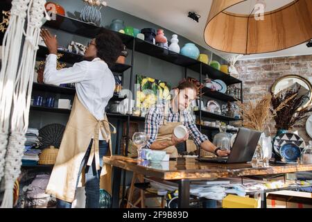 Two multicultural workers in uniform doing inventory together at decor shop. Bearded caucasian man using laptop while african woman counting goods. Stock Photo