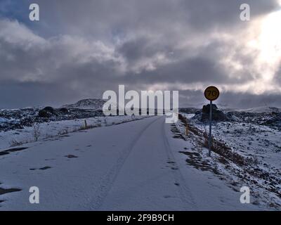 Diminishing perspective of snow-covered country road with tire marks and speed limit sign leading through lava field with rocks near Hafnarfjörður. Stock Photo
