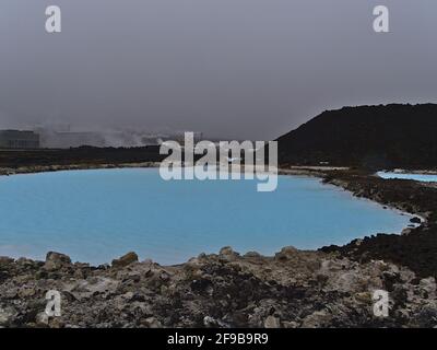 Pool with blue colored thermal water between lava rocks with steaming Svartsengi power station in background near Blue Lagoon, Grindavik, Iceland. Stock Photo