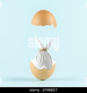 Easter egg wrapped in a paper in the shape of a bunny in cracked eggshell against pastel blue background. Easter minimal concept. Creative Happy Easte Stock Photo