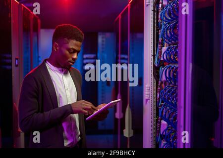 Serious young African-American data engineer standing at open server rack cabinet and using tablet while setting up system at data center Stock Photo