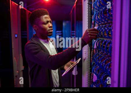 Concentrated young Black man using tablet while examining datacenter equipment in dark server room Stock Photo