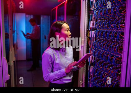 Concentrated young Asian woman in stripped shirt standing at server rack cabinet and using tablet while maintaining network in server room Stock Photo