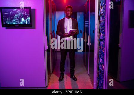 Serious young African-American network engineer in cardigan using tablet while checking connections at data center equipment Stock Photo