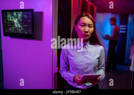 Portrait of serious Asian female network administrator standing with tablet against computer cabinets in server room Stock Photo