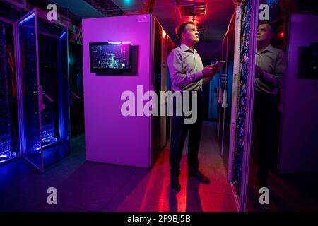 Concentrated male IT engineer standing in dark neon room full of server cabinets and using tablet while controlling network systems Stock Photo