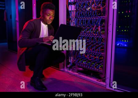 Concentrated young African American network engineer crouching by server cabinet and using laptop while examining work of supercomputer Stock Photo