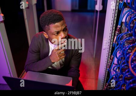 Thoughtful young black server engineer looking at cables in server cabinet while using laptop finding out error Stock Photo
