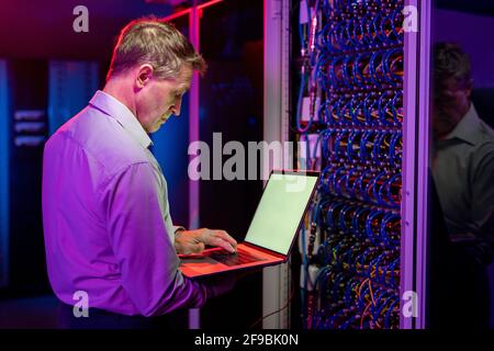 Serious mature network engineer standing at open server cabinet and using laptop while diagnosing server problems Stock Photo