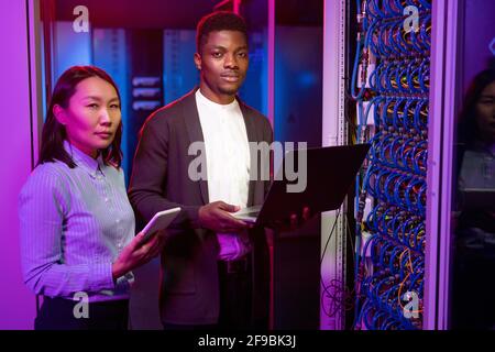 Portrait of confident multi-ethnic IT engineers standing with gadgets in dark blue server room and working with supercomputer Stock Photo