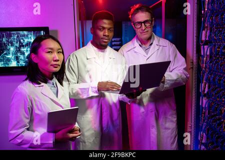 Portrait of confident multi-ethnic network scientists in lab coats standing with devices in neon-illuminated server room Stock Photo