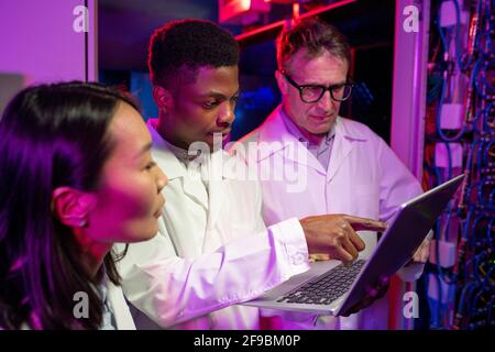 Group of concentrated modern multi-ethnic server specialists in lab coats using laptop while analyzing network error together in data center Stock Photo