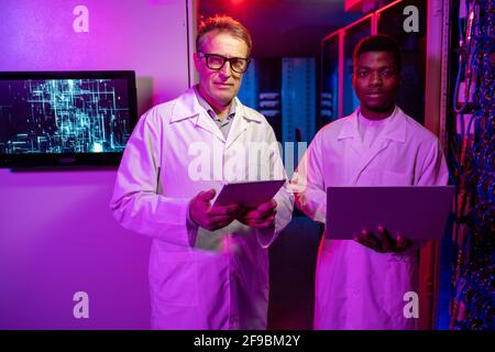 Portrait of multi-ethnic male server technicians in white coats standing with devices in dark neon-illuminated room with computer monitor on wall Stock Photo