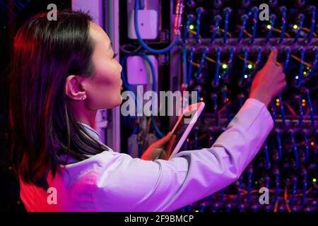 Rear view of busy Asian female network engineer in lab coat connecting cables in switches using tablet in dark room Stock Photo