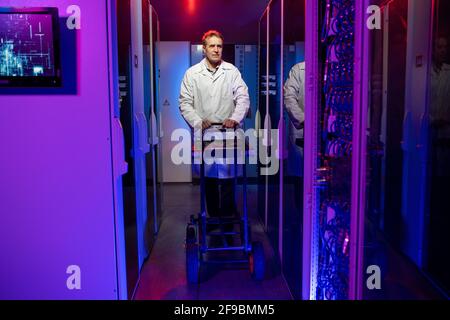 Mature server worker in white coat pushing cart with tools while walking over dark data center Stock Photo