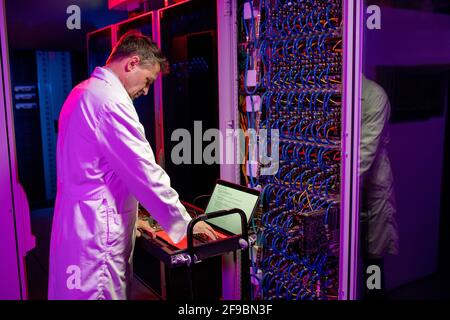 Concentrated mature network engineer in white coat standing at cart with laptop and configurating blade server in neon illuminated room Stock Photo