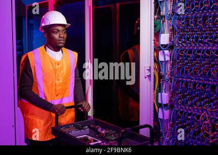 Serious young African-American repairman in hardhat and orange vest pushing cart with tools and devices and leaving server room Stock Photo