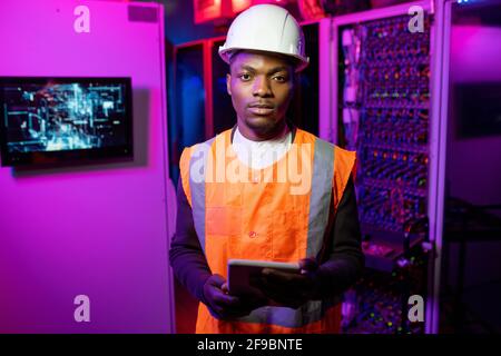Portrait of serious confident African American data center engineer in orange vest and white hardhat standing with tablet in server room in neon light Stock Photo