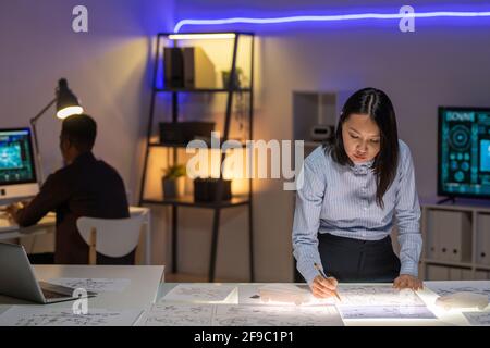 Concentrated young Asian designer in formal shirt standing at desk in dark neon office and drafting layout of car Stock Photo