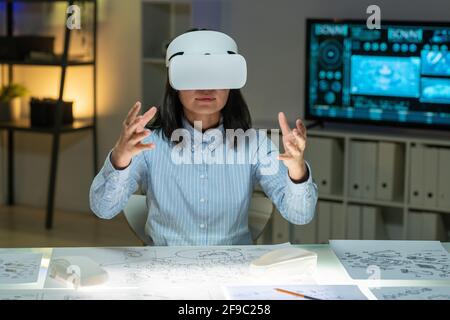 Content modern young Asian designer in stripped shirt sitting at table with layouts and gesturing hands while working in VR goggles Stock Photo