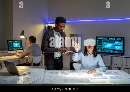 African American employee synchronizing tablet virtual reality headset of Asian colleague while they designing car in 3D Stock Photo