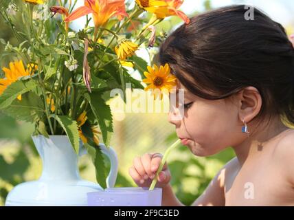 Funny little girl drinks water through a straw from a plastic cup while sitting at a table against the backdrop of sun-drenched vines Stock Photo