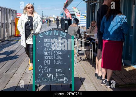 Southend on Sea, Essex, UK. 17th April, 2021. The day has been bright and sunny in the seaside town, but with a chill wind. Many people have visited to enjoy the seafront and its famous pier. The cafe at the enf of the pier has a humorous sign saying that cake is the answer to everything. Outside diners Stock Photo