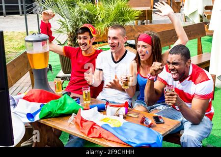 Young football supporter fans cheering with beer watching soccer match - Friends people with multicolored soccer tshirts and flags having fun Stock Photo