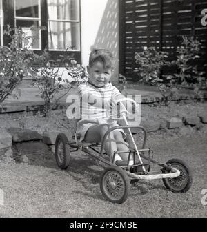 1966, historical, outside in a garden, a young boy, sitting, with his hands on the plastic steering wheel, in a four-wheeled metal-framed pedal toy car. As it is minus the outer shell - maybe broken over time - the toy could be a hand-med-down. Or looking at it, quite possibly a DIY home-made machine made from spare pieces of metal and four old pram wheels. Stock Photo