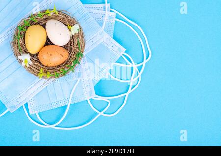 Nest with brightly dyed eggs with medical masks on blue background, space for text. Easter holiday during the Coronavirus pandemic. Studio Photo Stock Photo