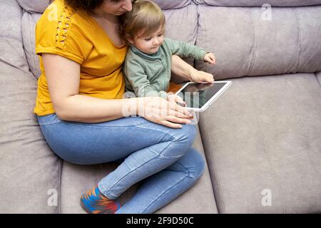 A young woman with her daughter sitting on the sofa at home using a tablet. Concept of family time, gadgets, conciliation, lifestyle. Stock Photo