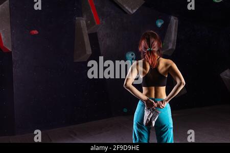 Young woman preparing to climb in bouldering gym, chalking hands and examining route on wall, rear view Stock Photo
