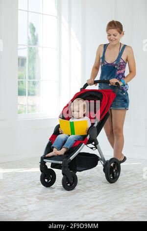 Caucasian young woman and toddler son in stroller indoors. Boy holding book and looking at pictures. Stock Photo