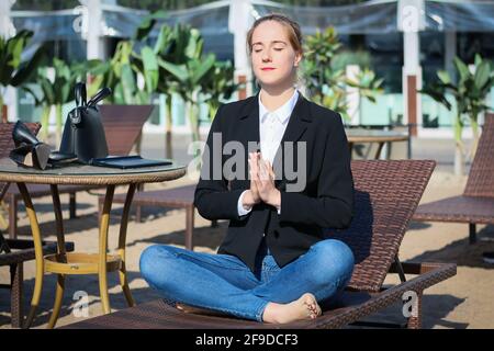 young beautiful business woman or freelancer takes coffee break at lounge and demonstrates meditation yoga pose sitting on deck chair  in summer sunny Stock Photo