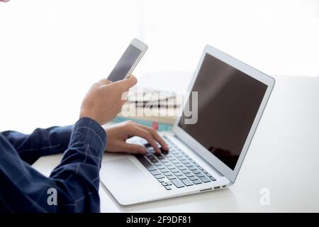 Hand of young man working with laptop computer and smartphone mockup on desk at home, notebook and phone display blank screen, freelance look message Stock Photo