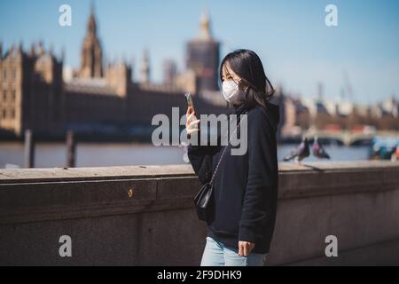 Westminster, London | UK -  2021.04.17: Young lady wearing medical mask taking pictures with phone at the Thames river embankment