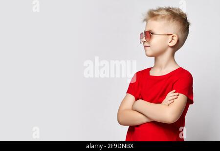 Portrait of blonde kid boy in red t-shirt and sunglasses standing with arms crossed at chest, looking at copy space Stock Photo