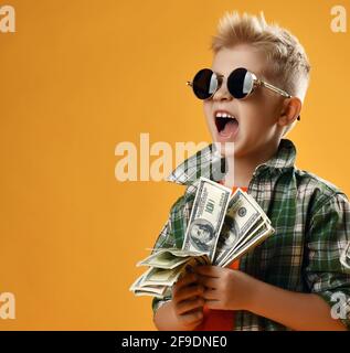 Blonde rich kid boy in round sunglasses and checkered plaid shirt stands with fan of dollar cash in hands and screams Stock Photo
