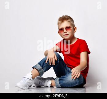 Cool blonde kid boy leader in red t-shirt, blue jeans, white sneakers and sunglasses sits on floor in relaxed pose Stock Photo