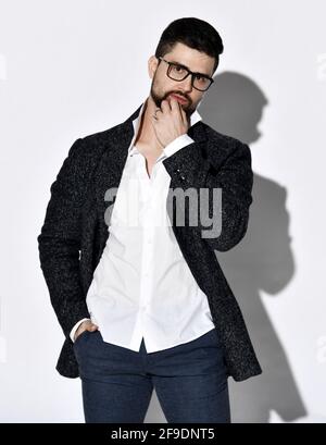 Pensive stylish bearded man in glasses, casual tweed jacket and jeans stands holding hand in his pocket and at his chin Stock Photo