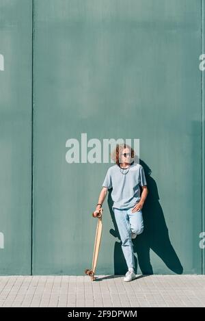 vertical portrait of a caucasian young man standing and holding his skateboard. He is wearing casual clothes and sunglasses. Green wall background Stock Photo