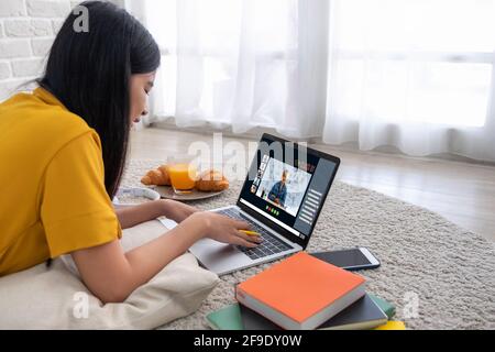 Asian female student video conference with teacher and friends using e-learing app on laptop computer at home.woman lying down on carpet.new normal le