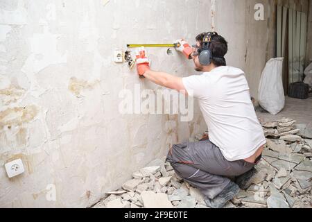 Builder man using a measuring tape at the construction site. Construction worker wearing safety gloves, glasses and earmuffs. Stock Photo