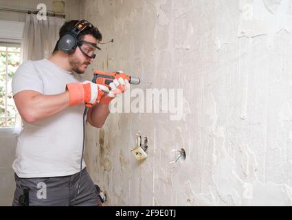 Builder drilling holes in a wall with an electric drill at the construction site. Construction worker wearing safety gloves, glasses and earmuffs. Stock Photo