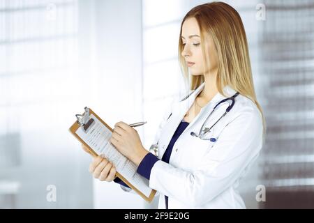 Young woman-doctor is making some notes using a clipboard, while standing in her cabinet in a clinic. Portrait of beautiful female physician with a Stock Photo