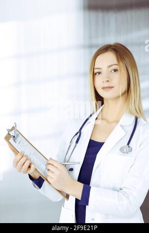 Young intelligent woman-doctor is prescribing some medicine for a patient, using a clipboard, while standing in the cabinet in a clinic. Female Stock Photo