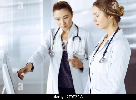 Two female physicians are discussing medical therapy, while standing at the table in a clinic office. Doctors use pc computer at work. Teamwork in Stock Photo